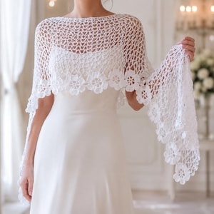 Puede incluir: Un chal de ganchillo blanco con un intrincado patrón de malla calada en los hombros y la parte superior de la espalda, rematado con un borde decorativo de motivos florales repetidos a lo largo del bajo, sobre un vestido blanco sin mangas.