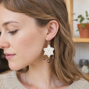 May include: White crochet star-shaped earrings with gold-colored hooks and three dangling brown beads. The earrings are handmade and have a delicate, open weave design. The earrings are a unique accessory.