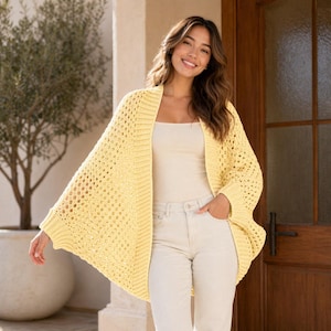 May include: A woman wearing a pale yellow, open-front crochet cardigan. The cardigan features a loose, flowing design with an open weave. She is also wearing a white vest top and light-coloured jeans. The background includes a tree and a wooden door.