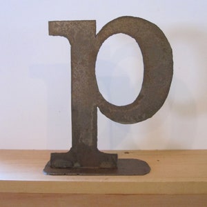 May include: A rusty metal letter P, standing on a wooden shelf. The letter is made of thick metal and has a rustic, industrial look.