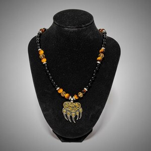 May include: A necklace with a bear claw pendant, displayed on a black mannequin. The necklace features black and amber-coloured beads, with silver accents. The pendant has a gold-coloured Celtic knot design.
