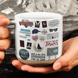 Peut inclure: Mug en céramique blanche avec une anse grise et une base marron, décoré de divers autocollants et citations. Les autocollants incluent un camion rouge, des lunettes de soleil et les phrases "Do I dazzle you ?" et "Let's start with Forever."