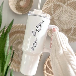 May include: A white stainless steel tumbler with a handle and a clear lid featuring a straw. The tumbler is decorated with two identical black and white stickers, each depicting a cartoon microscope positioned above a small, smiling potato character.