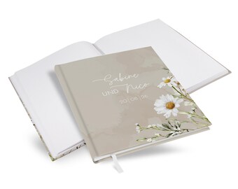 May include: A hardcover guest book with a beige cover featuring white daisy illustrations and the printed text Sabine UND Nico 20 | 08 | 26. The book is shown open, revealing blank white pages and a small white ribbon bookmark attached to the spine.