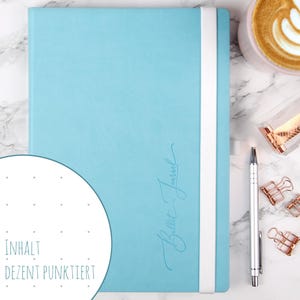 Può includere: Un diario azzurro con una fascia elastica bianca di chiusura. La copertina presenta il testo Bullet Journal in corsivo. Un inserto circolare mostra una pagina bianca con una griglia di punti e il testo INHALT DEZENT PUNKT.