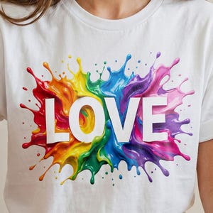 May include: White t-shirt with a vibrant paint splash design. The splash is in rainbow colours and surrounds the word "LOVE" in white capital letters. The shirt is short-sleeved.