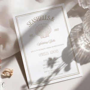 May include: A rectangular white invitation card with a gold border featuring a central seashell illustration. The text reads: SEASHELLS & 2021, EST., Wedding Bells, JOIN US TO CELEBRATE A BRIDAL SHOWER HONORING OPHELIA ADAMS, JUNE 26 4 PM, THE ADAM'S RESIDENCE.