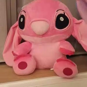 Puede incluir: Un personaje de peluche rosa con grandes ojos negros, orejas largas y una nariz peque&ntilde;a. El peluche tiene un cuerpo redondo, con c&iacute;rculos rojos en los pies y las manos. El juguete est&aacute; sobre una superficie blanca.