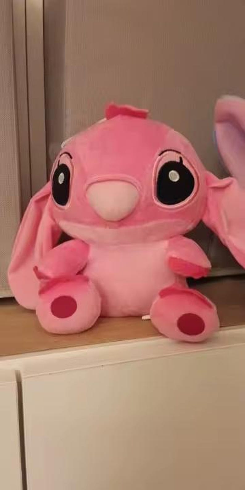 Puede incluir: Un personaje de peluche rosa con grandes ojos negros, orejas largas y una nariz peque&ntilde;a. El peluche tiene un cuerpo redondo, con c&iacute;rculos rojos en los pies y las manos. El juguete est&aacute; sobre una superficie blanca.