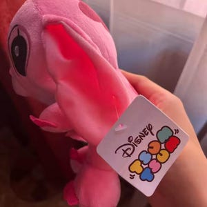 Puede incluir: Un peluche rosa de un personaje con orejas grandes y una etiqueta que dice "Disney Tsum Tsum". El juguete es de color rosa claro con manchas y detalles en rosa m&aacute;s oscuro. La etiqueta es blanca con logotipos coloridos de Disney.