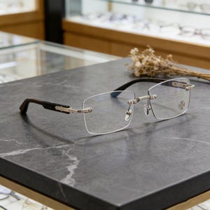 May include: Rimless eyeglasses with rectangular lenses. The frames feature a silver metal bridge and hinges, with dark brown temples. A small decorative emblem is on the right lens. The glasses rest on a dark gray marble surface.