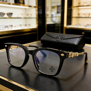 Puede incluir: Gafas negras de montura cuadrada con detalles dorados y un emblema de cruz decorativo en las lentes. Las patillas presentan detalles dorados ornamentados y un pequeño emblema dorado en el extremo. Un estuche de cuero negro está en el fondo.