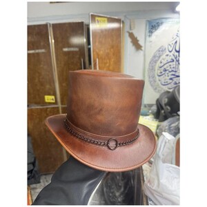 Puede incluir: Un sombrero de copa marrón con una textura suave similar al cuero. El sombrero presenta una banda de cadena de metal oscuro envuelta alrededor de la base de la copa, que incluye un pequeño detalle de anillo de metal circular en el frente.