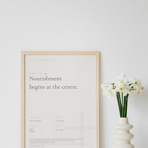 Puede incluir: Un estampado enmarcado con el texto "Nourishment begins at the centre." en un marco de madera clara. Un jarrón blanco con un diseño único contiene flores blancas con centros amarillos. El tema general es minimalista y relajante.