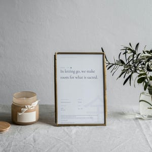 Puede incluir: Una impresión enmarcada con el texto "In letting go, we make room for what is sacred" se encuentra junto a una vela encendida en un tarro de cristal con tapa de madera. Un pequeño jarrón de cristal contiene ramas de olivo.