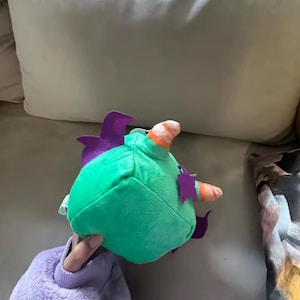 May include: A plush toy in the shape of a green creature with purple fins and a striped horn. The toy is held in a person's hand, and the background is a light-colored couch.