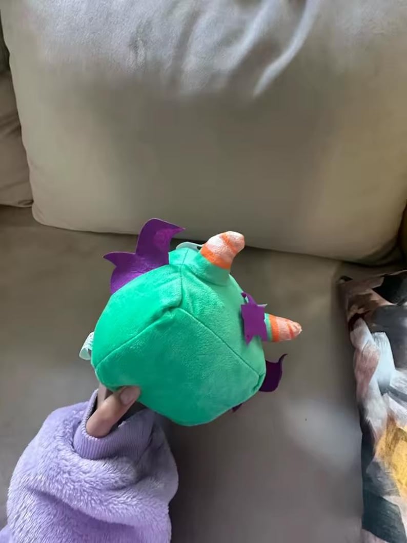 May include: A plush toy in the shape of a green creature with purple fins and a striped horn. The toy is held in a person's hand, and the background is a light-colored couch.