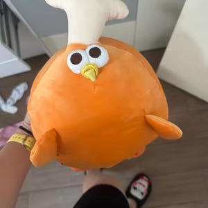 May include: An orange plush toy shaped like a cartoon character with a white head crest, large eyes, and a yellow beak. The toy has small orange fins and is held by a person.