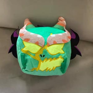 May include: A plush toy in the shape of a monster's head. The toy is primarily green with yellow, orange, and black accents. It has large yellow eyes, orange horns, and black wing-like shapes on the sides.