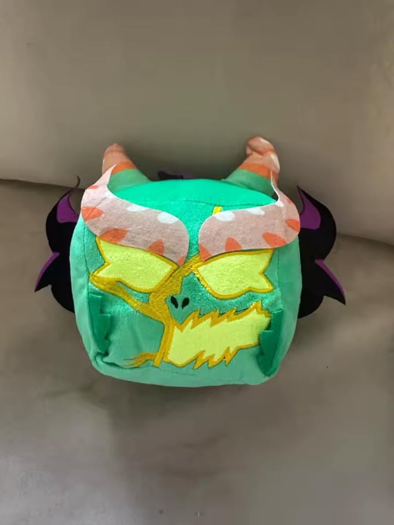 May include: A plush toy in the shape of a monster's head. The toy is primarily green with yellow, orange, and black accents. It has large yellow eyes, orange horns, and black wing-like shapes on the sides.