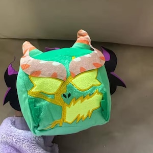 May include: A green plush toy shaped like a monster's head with yellow embroidered details, orange and white horns, and purple wings. The toy is held up against a neutral background.