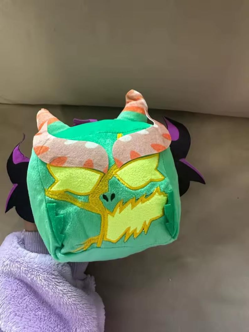 May include: A green plush toy shaped like a monster's head with yellow embroidered details, orange and white horns, and purple wings. The toy is held up against a neutral background.