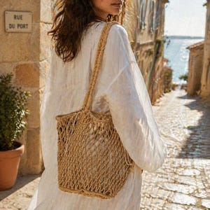 May include: A woven beige tote bag with a braided handle is carried by a person wearing a white linen shirt. The bag has a net-like design. The background shows a cobblestone street and a sign that reads "RUE DU PORT."