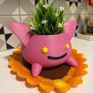 May include: A pink, rounded planter with two pointed ears and two small feet, featuring two yellow circular eyes and a black curved mouth. The planter sits on a flat, orange and brown sunflower-shaped base and contains green grass with small white flowers.