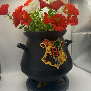 Puede incluir: Un jarrón negro en forma de caldero con un escudo de Hogwarts, lleno de flores artificiales rojas y blancas y follaje verde. El jarrón tiene dos asas y se asienta sobre una base gris. Las flores tienen centros amarillos.