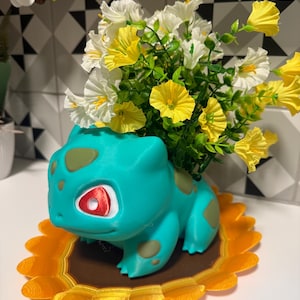 May include: A teal creature-shaped planter with olive green spots and red eyes, containing a bouquet of artificial white and yellow flowers. The planter sits on a brown and orange scalloped base that resembles a sunflower with yellow petals.
