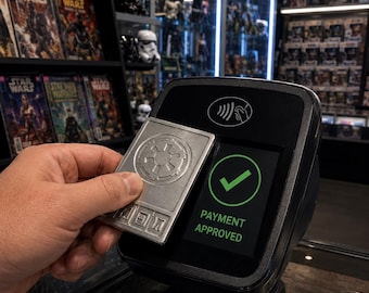 May include: A rectangular silver-colored card featuring an embossed Imperial crest from Star Wars being held against a black contactless payment terminal. The terminal screen displays a green checkmark icon and the text PAYMENT APPROVED.