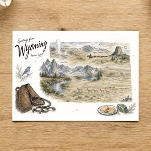 May include: A rectangular postcard featuring a watercolour illustration of Wyoming with the text Greetings from Wyoming Forever West. The map depicts mountains, wildlife, and landmarks like Old Faithful and Devils Tower, alongside leather chaps, spurs, a blue bird, and biscuits.