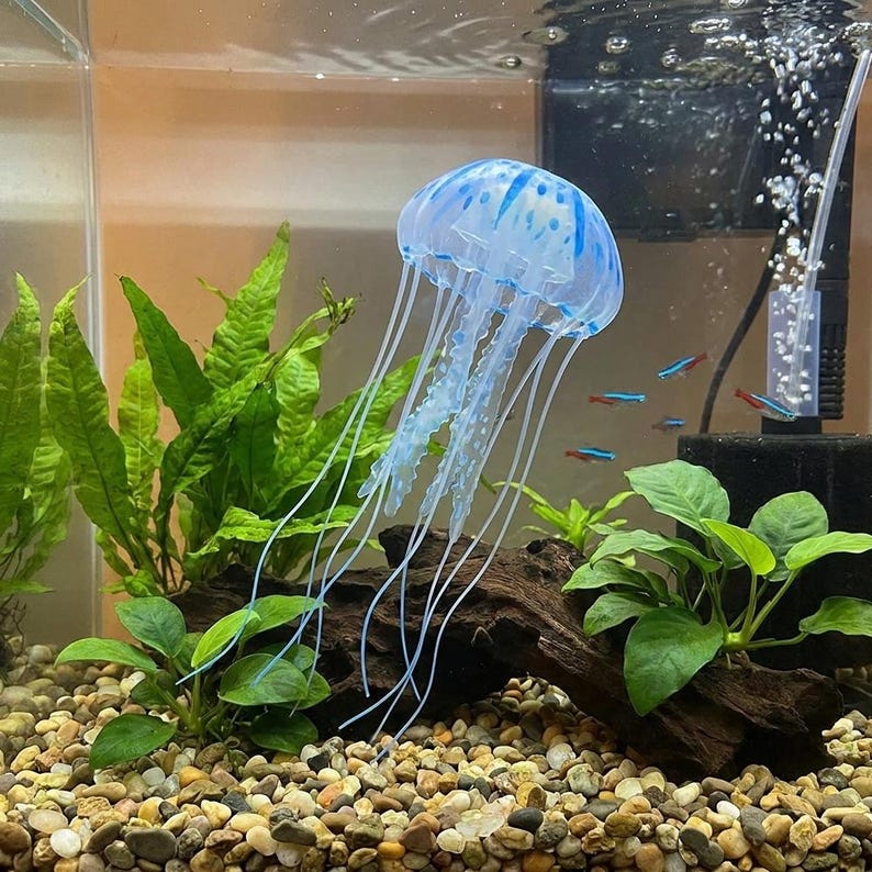 Pu&ograve; includere: Una decorazione a medusa blu e bianca galleggia in un acquario. La medusa ha lunghi tentacoli fluenti ed &egrave; circondata da piante verdi, piccoli pesci e un substrato di ghiaia. L'acquario ha anche un filtro e una pompa d'aria.