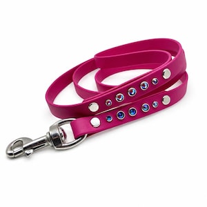 May include: A hot pink dog lead featuring a smooth, flexible strap with a silver-toned metal swivel snap hook. The lead is embellished with a series of circular silver studs and small, iridescent blue crystals arranged in a linear pattern.