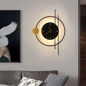 Puede incluir: Reloj de pared moderno con esfera negra, manecillas doradas y un acento dorado. El reloj está dentro de un marco circular negro con una luz detrás. Dos líneas verticales negras y un círculo dorado completan el diseño.