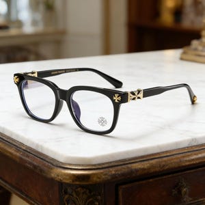 May include: Black-framed eyeglasses with gold detailing. The arms and hinges have gold cross motifs. The lenses are clear, and the frame is a glossy black. The glasses are presented on a white marble surface.