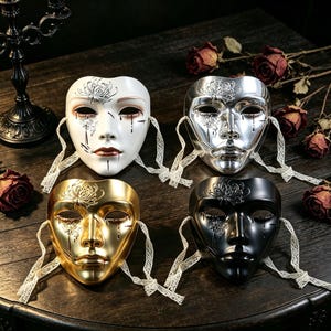 Op de afbeelding: Vier maskers in wit, zilver, goud en zwart, elk voorzien van een reliëf bloemmotief van een spinnenlelie op het voorhoofd en de wang, geschilderde traanvormige accenten en witte kanten linten aan de zijkanten om het masker vast te maken.
