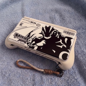 May include: A white Nintendo 3DS console with a black and white monster graphic and Japanese text. The handheld device has a wrist strap with a brown and beige cord. The console is a handheld gaming device.