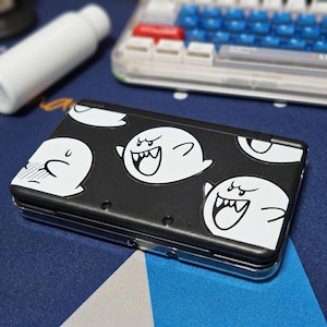 May include: A black handheld gaming console featuring a repeating white graphic pattern of Boo ghosts with various expressions. The device has a smooth matte finish and is enclosed in a clear protective plastic shell, showcasing the detailed character art.