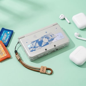 Pode incluir: Um console de jogos portátil cinza claro com uma capa protetora transparente com um design de personagem de anime azul. O console tem uma pulseira de pulso combinando. Fones de ouvido sem fio brancos e estojos de carregamento também são visíveis.