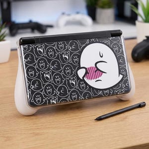 Puede incluir: Una Nintendo Switch Lite en blanco y negro con una funda protectora con un diseño de fantasma de dibujos animados. La funda tiene agarres blancos y un lápiz negro está al lado, sobre una superficie de madera.