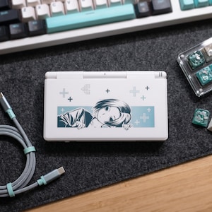 May include: A white handheld gaming console featuring a graphic of an anime-style character with short hair resting their chin on their hands. The design includes a light blue rectangular background with small plus signs and dotted arrow patterns.