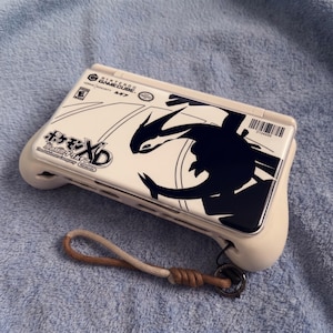 Puede incluir: Una consola portátil Nintendo GameCube blanca con un gráfico negro de una criatura con forma de dragón. La consola tiene un agarre color crema y un cordón. El texto "Pokémon XD" es visible.