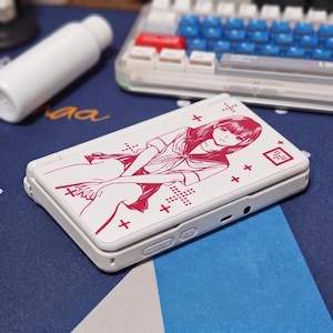 May include: A white handheld gaming console featuring a magenta-coloured line art illustration of a person with long hair wearing a collared shirt. The design includes small cross symbols and a square icon with a dot pattern on the top surface.