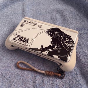 Könnte beinhalten: Ein weißer Nintendo GameCube-Controller mit einer schwarz-weißen Grafik von Link aus The Legend of Zelda: Twilight Princess. Inklusive passender Handschlaufe.