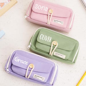 May include: Three rectangular pencil cases in pastel pink, sage green, and lavender. Each case features a front flap with a white button closure, a braided cord loop, and a small rectangular label reading "my study accept". Names "Sofia", "Oliver", and "Grace" are printed in white on the flaps.