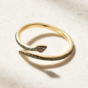 May include: A gold-toned wrap ring shaped like a snake, featuring a series of small green stones embedded along the band and two small orange stones set within the head of the snake, with a smooth, polished finish on the inner surface of the band.