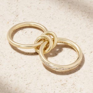 May include: A gold-colored ring set featuring three interconnected bands. One band is adorned with small, rectangular gemstones. The rings are displayed on a light-colored surface, showcasing their polished finish and intricate design.