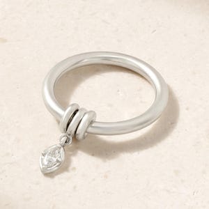 Puede incluir: Un anillo de tono plateado con una banda lisa y redondeada que incluye dos pequeños anillos móviles. Una piedra preciosa transparente de talla marquesa cuelga de estos anillos, creando un efecto de dije colgante en la banda principal.