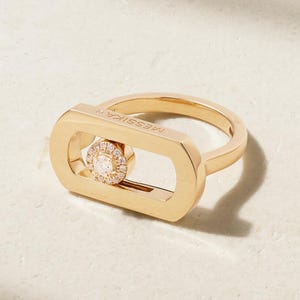 Könnte beinhalten: Ein goldener Ring mit einem rechteckigen Rahmen mit abgerundeten Kanten und dem eingravierten Schriftzug MESSIKA auf dem oberen Steg. Ein kreisförmiger, mit Diamanten besetzter Anhänger ist in der Mitte der rechteckigen Öffnung beweglich.
